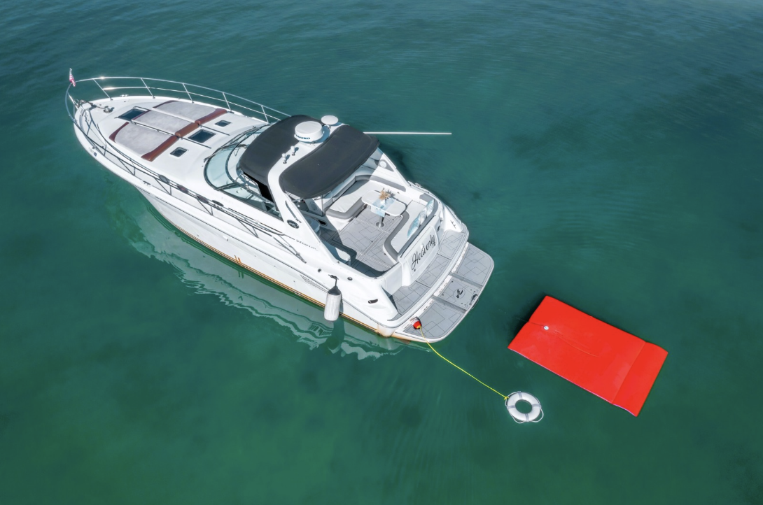 Aerial view of a white yacht anchored on turquoise water with a red floating mat attached by a rope.