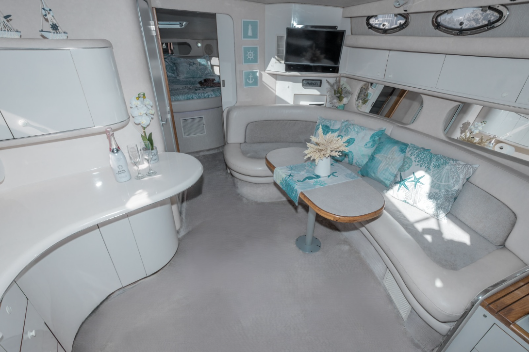 Luxury yacht interior with curved sofa, dining table, and television. Elegant decor includes decorative pillows and a floral arrangement.