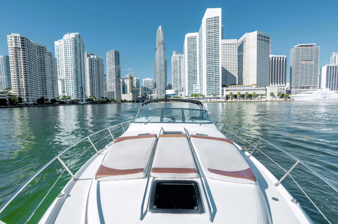 View from a boat on water facing a city skyline with tall modern buildings under a clear blue sky.