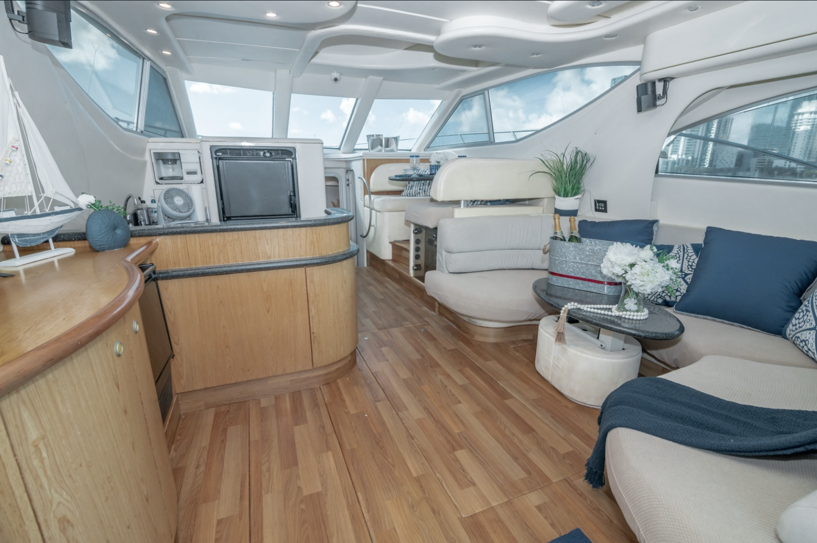Luxury yacht interior with wood flooring, plush seating, decorative cushions, potted plant, mini fan, sailboat model, and a table with champagne.