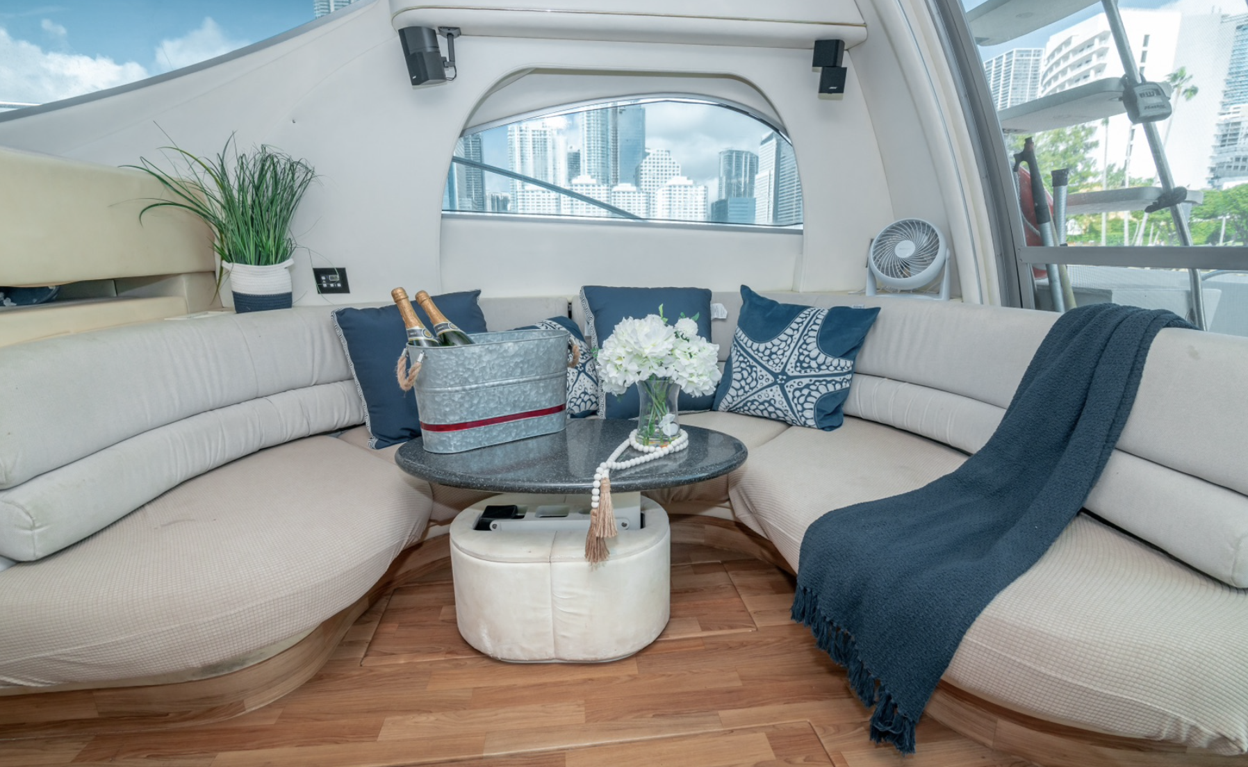 Luxury yacht interior with curved beige sofa, blue cushions, a metal ice bucket with champagne bottles, and a table with flowers.