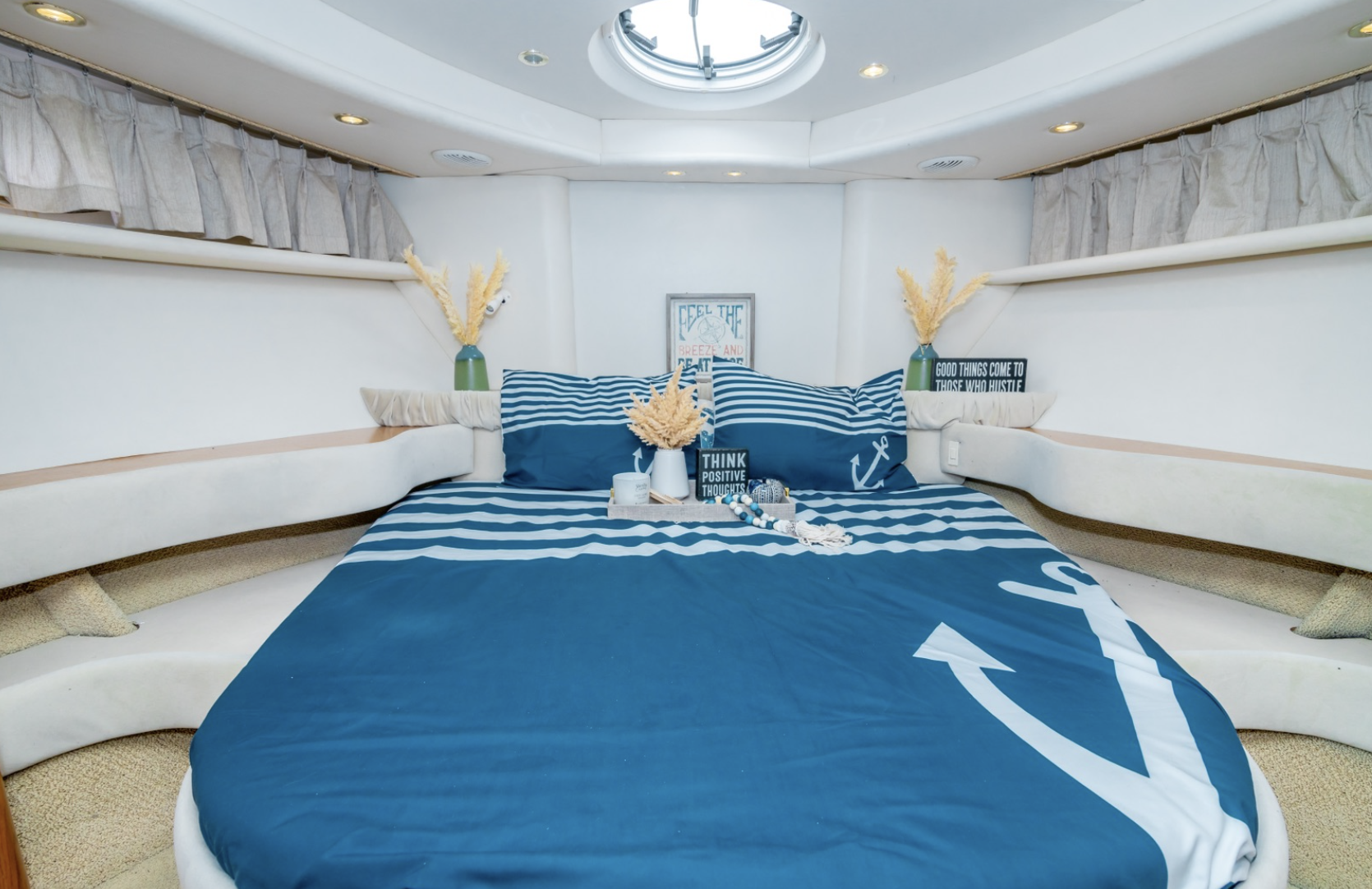 Cozy yacht bedroom with nautical theme bedding, featuring an anchor design. Decor includes pillows, vases with pampas grass, candles, and motivational signs. Circular ceiling window provides light.