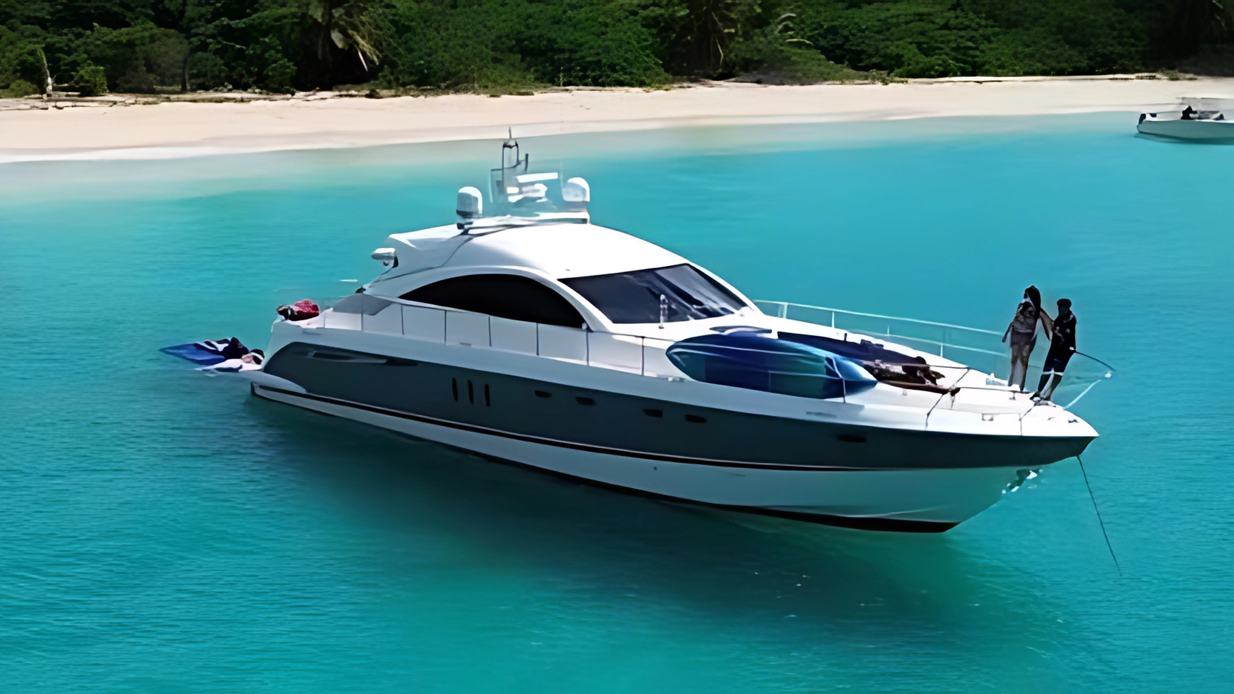 Luxury white yacht anchored in turquoise water near a sandy beach with green trees in the background, with two people standing on the bow, and a small boat tied to the side.