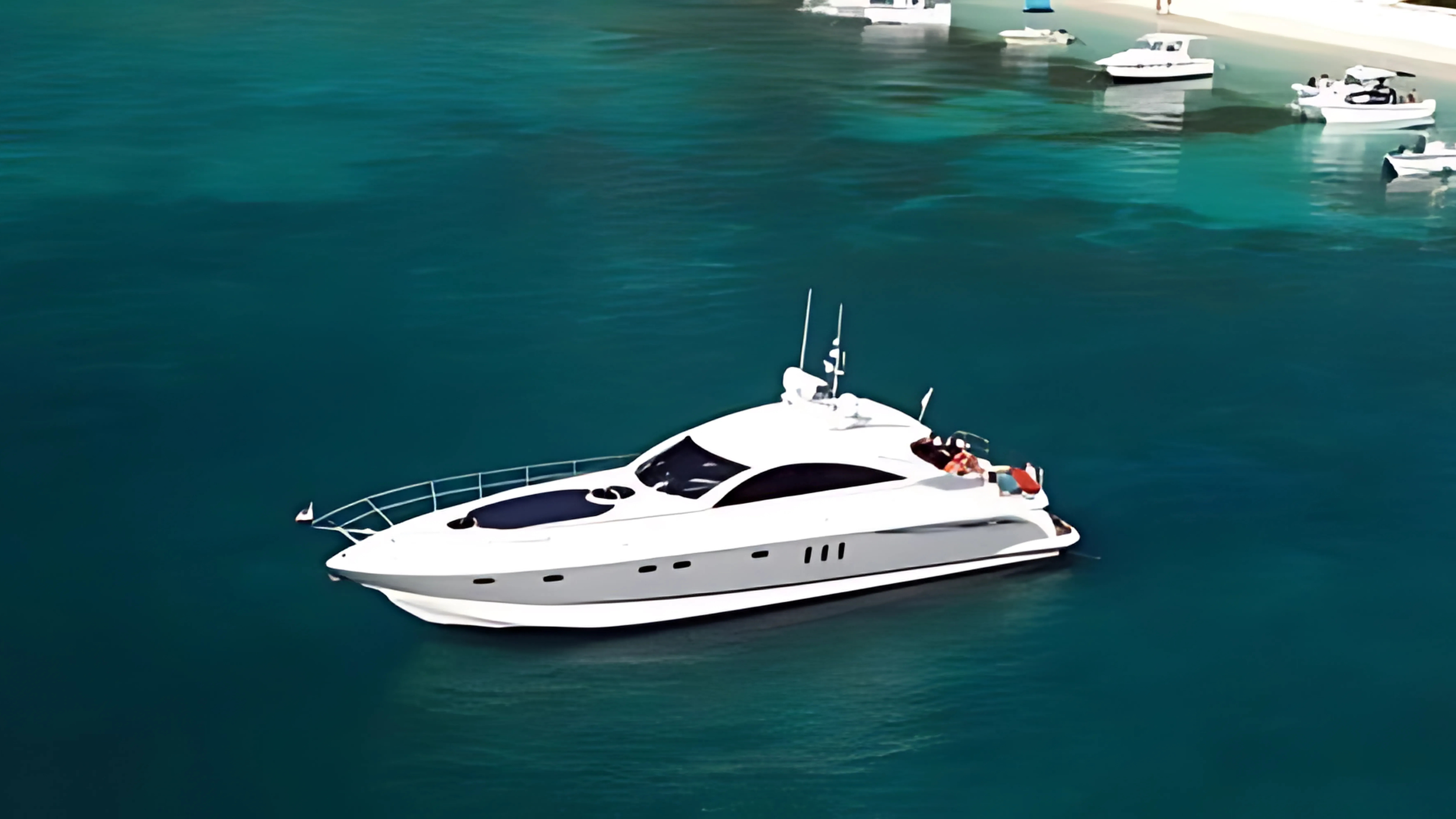 A white luxury yacht floating on calm blue-green water near a dock with multiple small boats moored in the background.