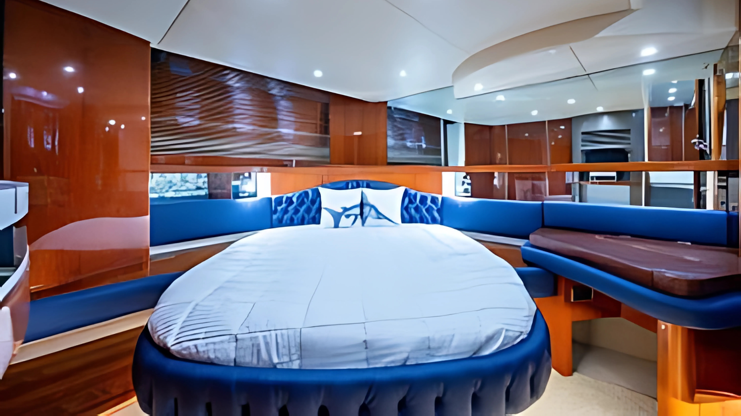 Interior of a luxurious yacht bedroom with a large, round bed, blue upholstered headboard, and wooden furniture and paneling.