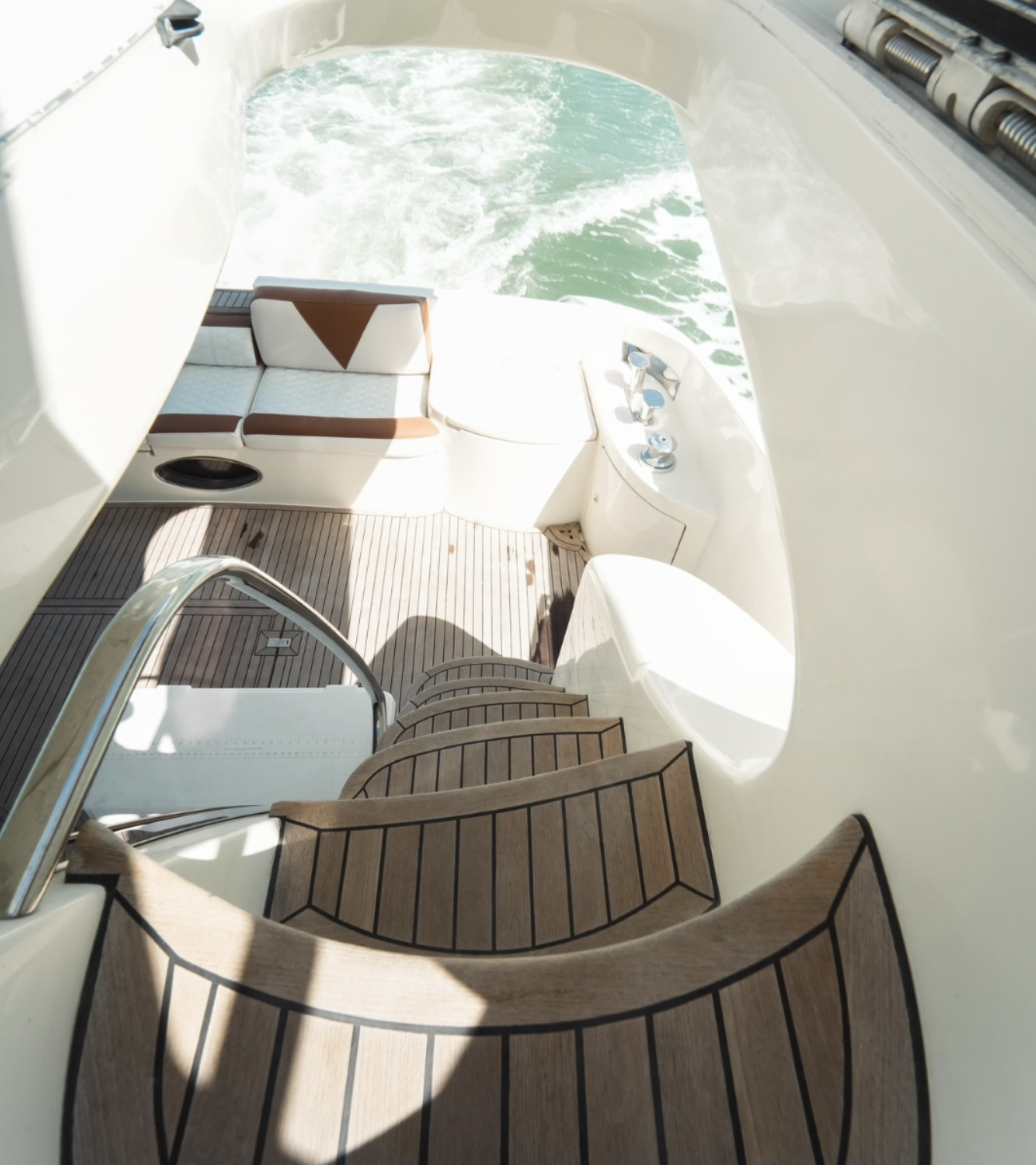 View from the upper deck of a yacht showing a curved wooden staircase, a cushioned seating area, and control panels with water in the background.