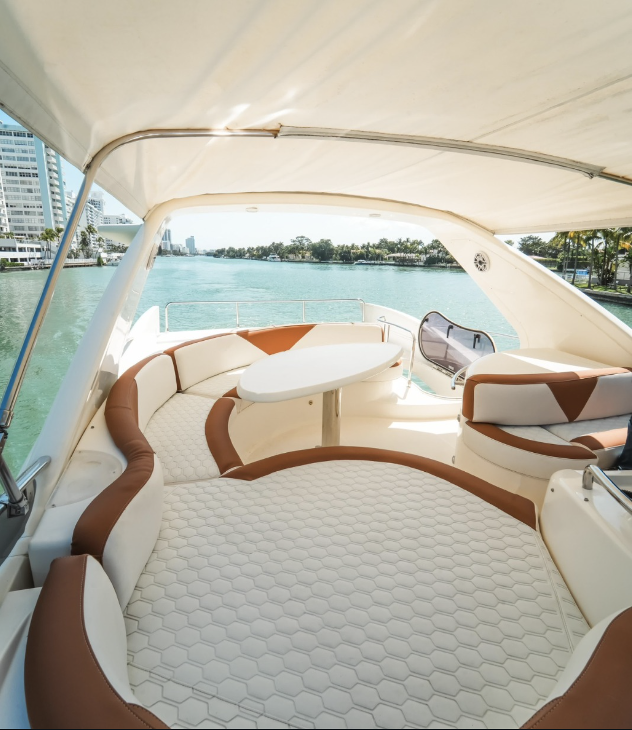 Interior of a luxury yacht with cushioned seating, a small table, and a cover over the deck, overlooking water and cityscape in the background.
