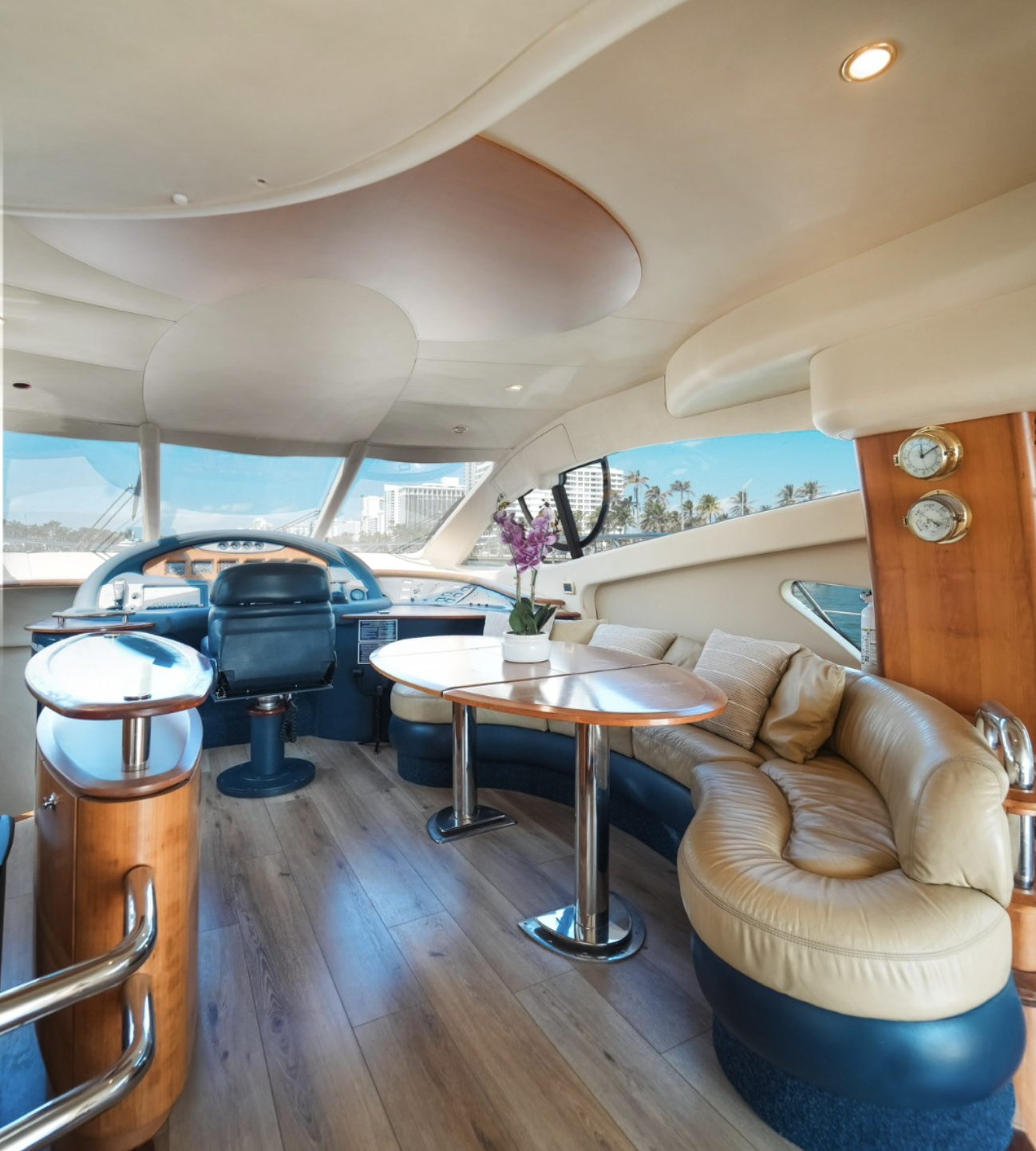 Interior of a yacht with a beige leather curved sofa, a wooden table with a purple orchid, and a ship's control area with a black leather chair, located near large windows showing a city skyline and palm trees outside.