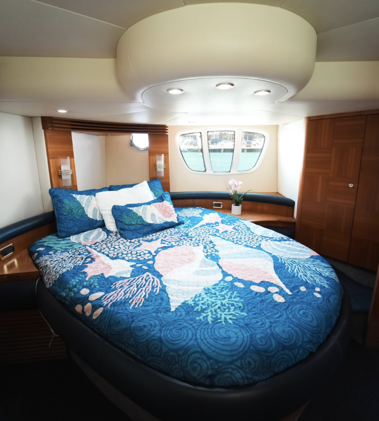 Interior of a boat bedroom with a round bed, Ocean-themed bedding, wooden accents, a small porthole window, and a larger boat window showing water and other boats outside.