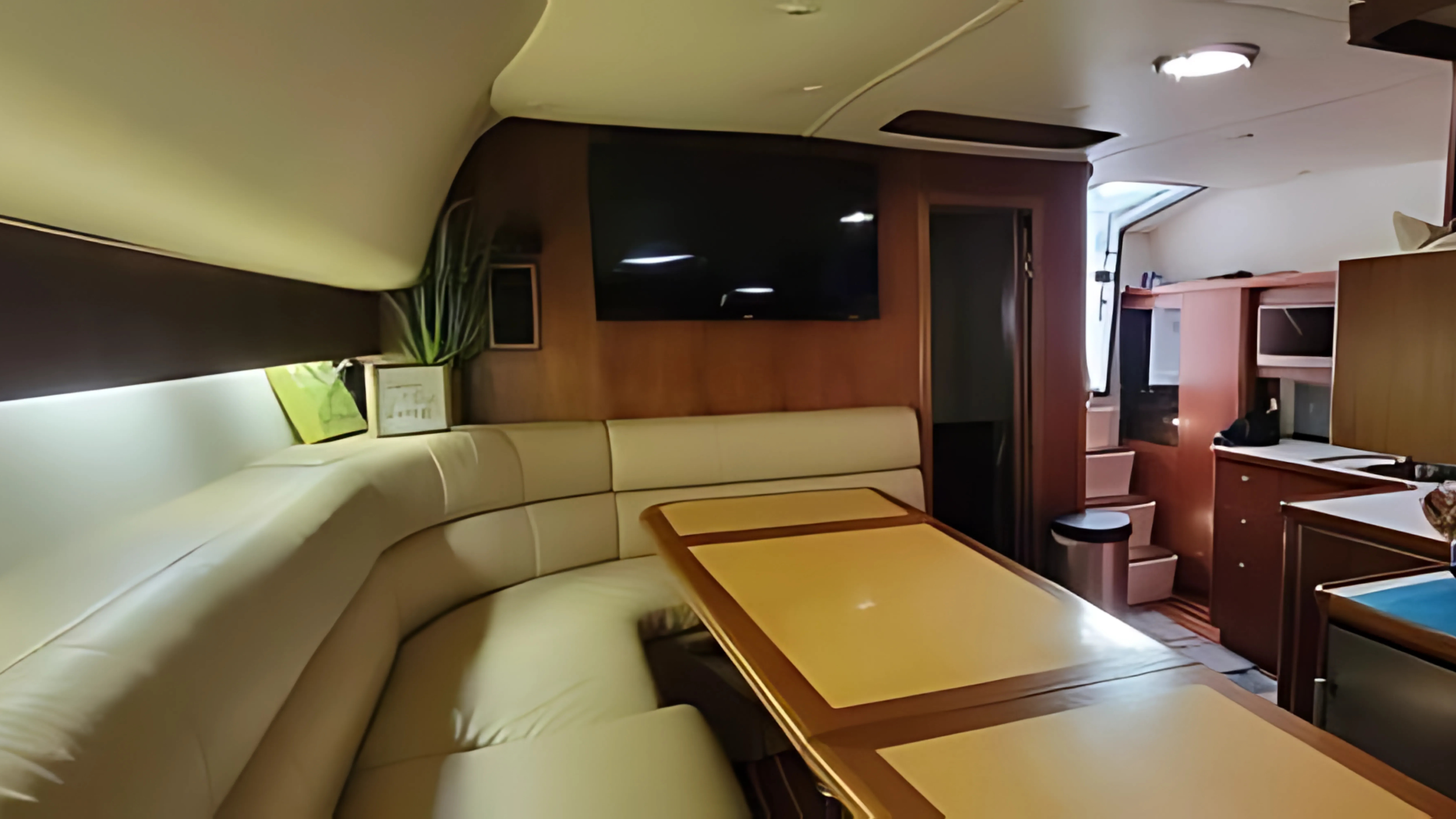 Interior of a small room with a cream curved sofa, a wooden table, a wall-mounted TV, a potted plant, a window, and various furniture and appliances.