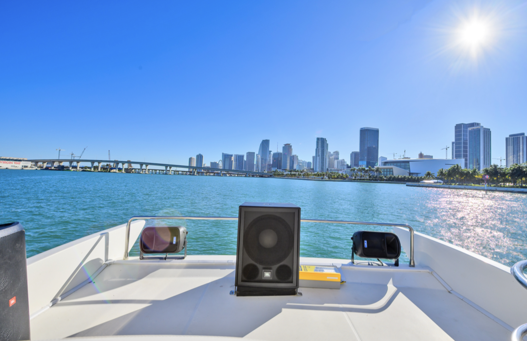 View from a yacht deck of Miami skyline across Biscayne Bay with blue sky and sun, featuring tall buildings and a bridge, and speakers on the deck.