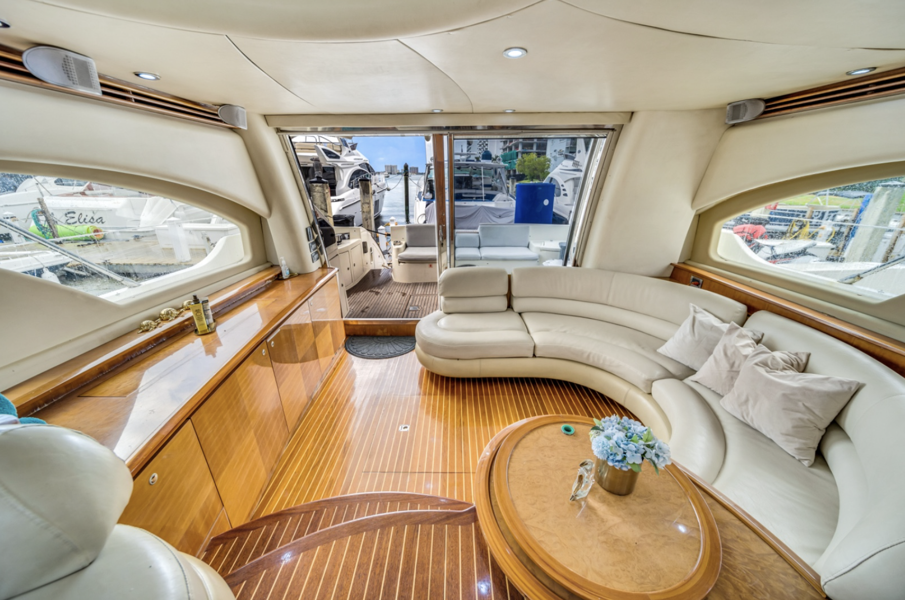 Luxury yacht interior with white leather seating, wooden flooring, a coffee table with flowers, and large windows overlooking a marina.
