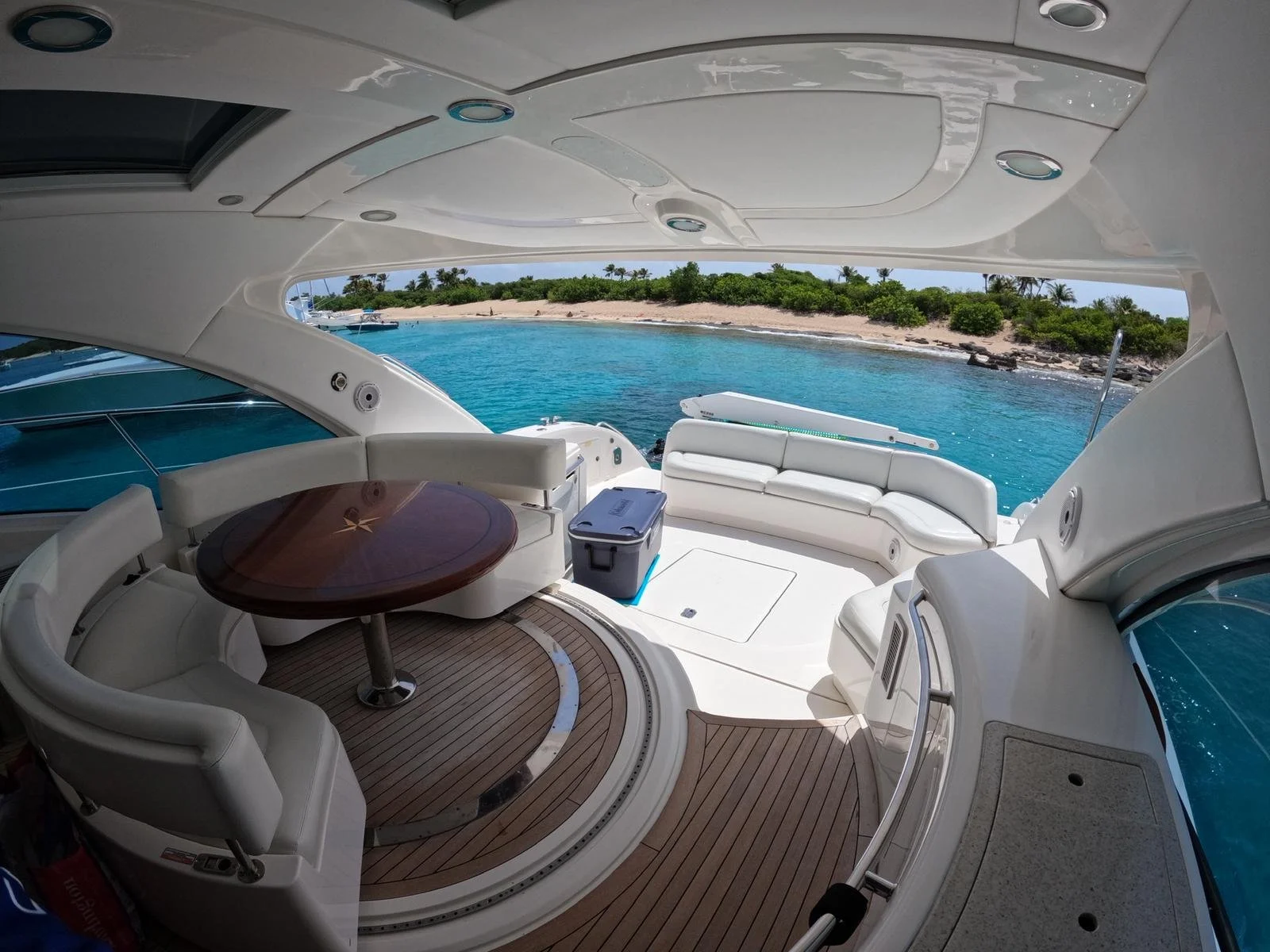 Interior of a luxury yacht with a wooden table and white leather seating, overlooking a tropical beach with clear blue water and palm trees.