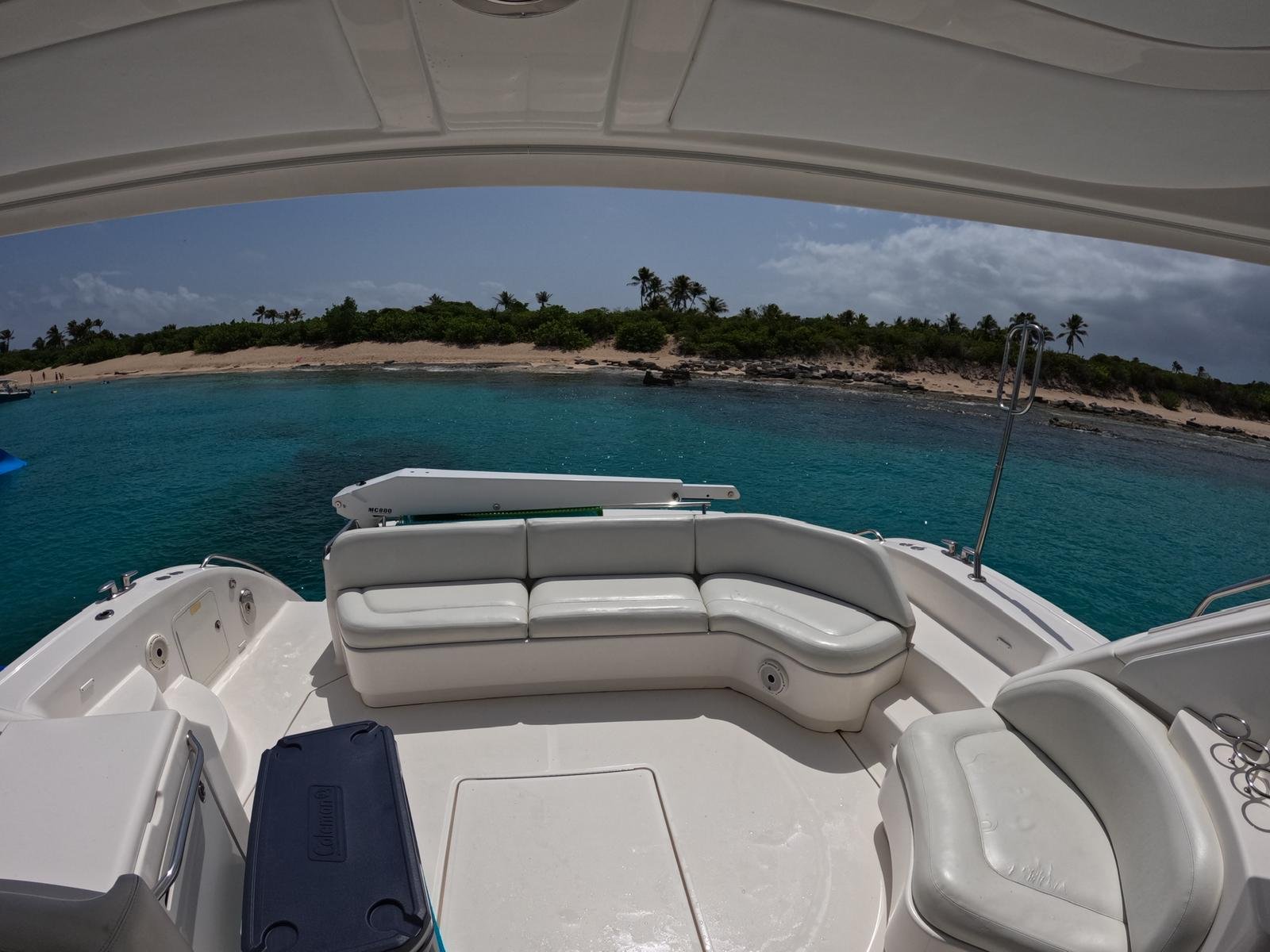 View from a boat with white seating and a cooler, overlooking a tropical beach with palm trees and clear blue water.