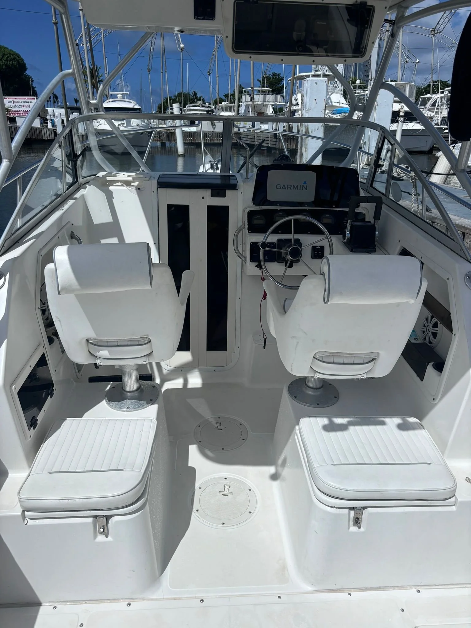 Interior of a yacht cockpit featuring two white captain chairs, a steering wheel, and a Garmin navigation system, with a marina in the background.