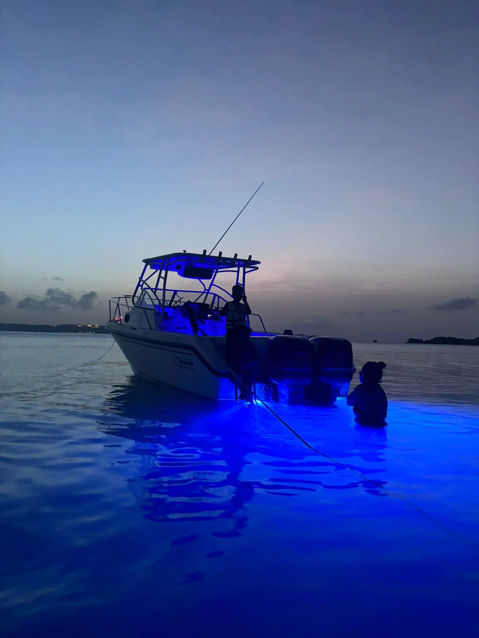 Boat illuminated with blue lights on calm water at dusk, person standing nearby.