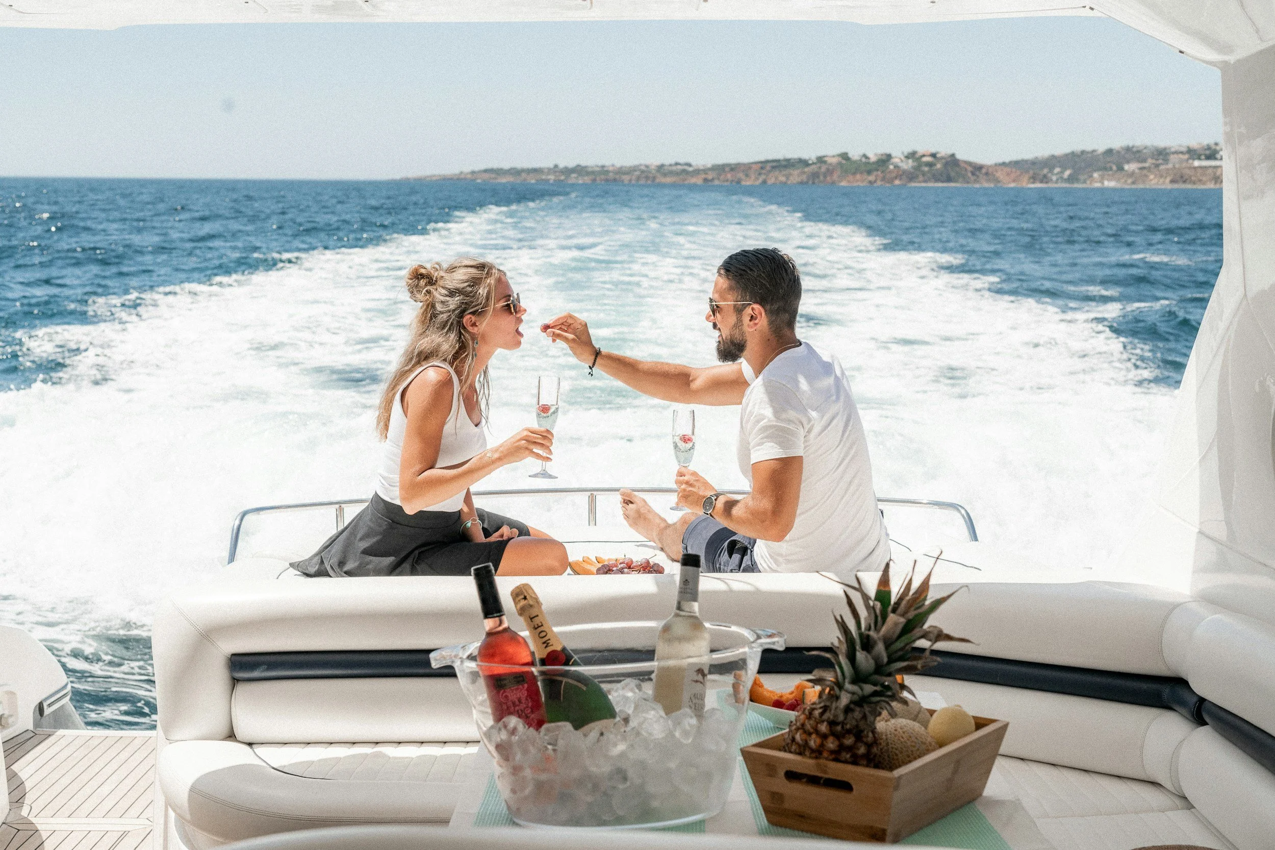 A couple on a yacht enjoying drinks and music, with the ocean and a distant landmass in the background.