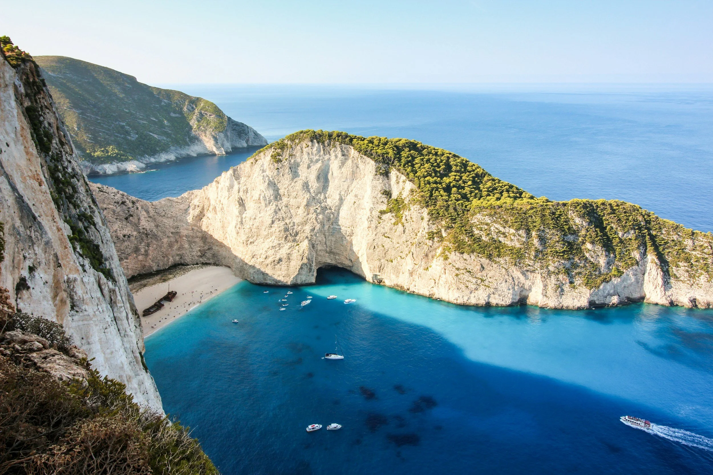 A scenic aerial view of a coastal landscape featuring white chalk cliffs, a sandy beach, turquoise water, boats, and lush green vegetation on the cliffs.