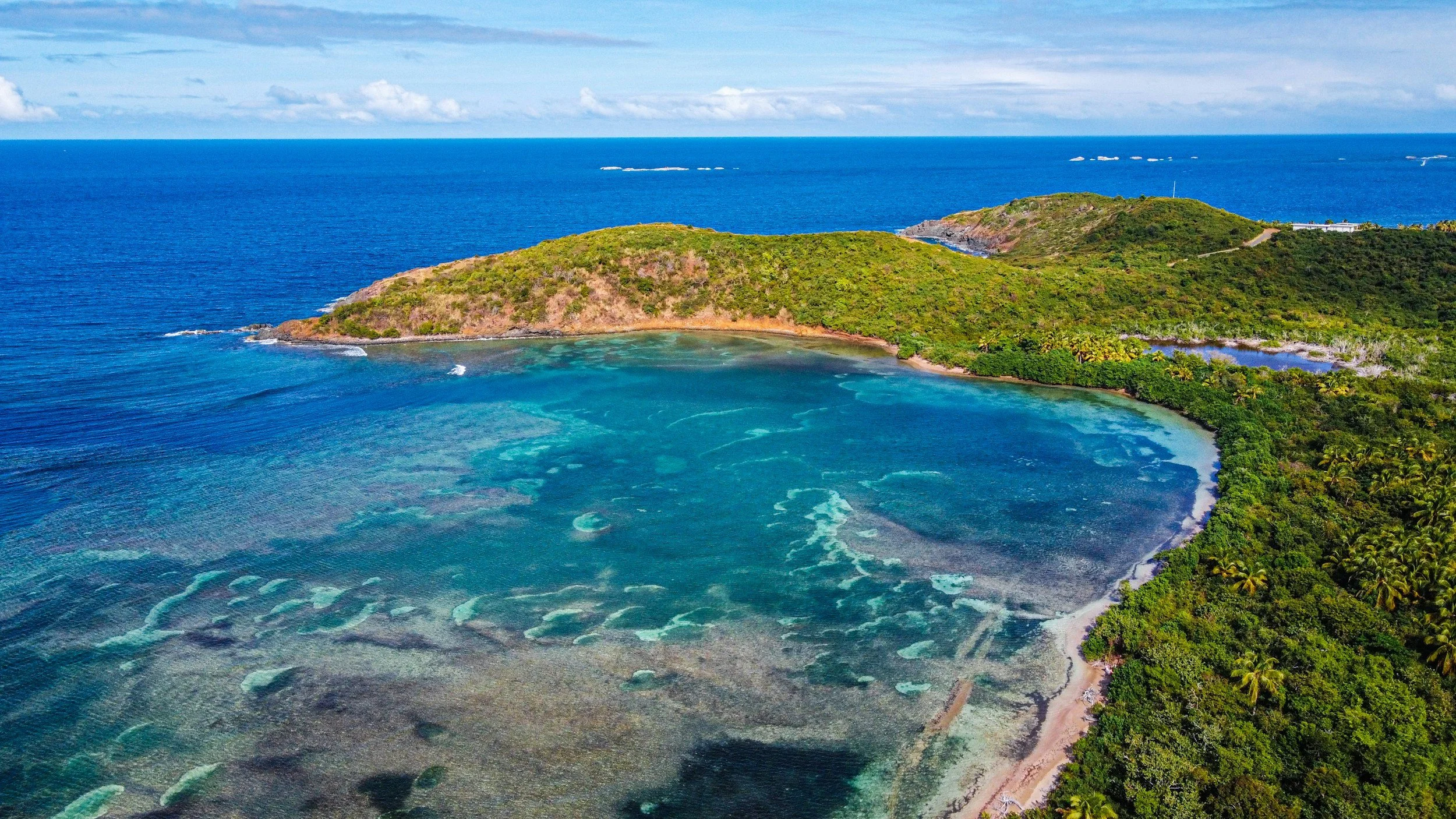 Aerial view of a coastal landscape with a bay, green hills, and the ocean in the background.