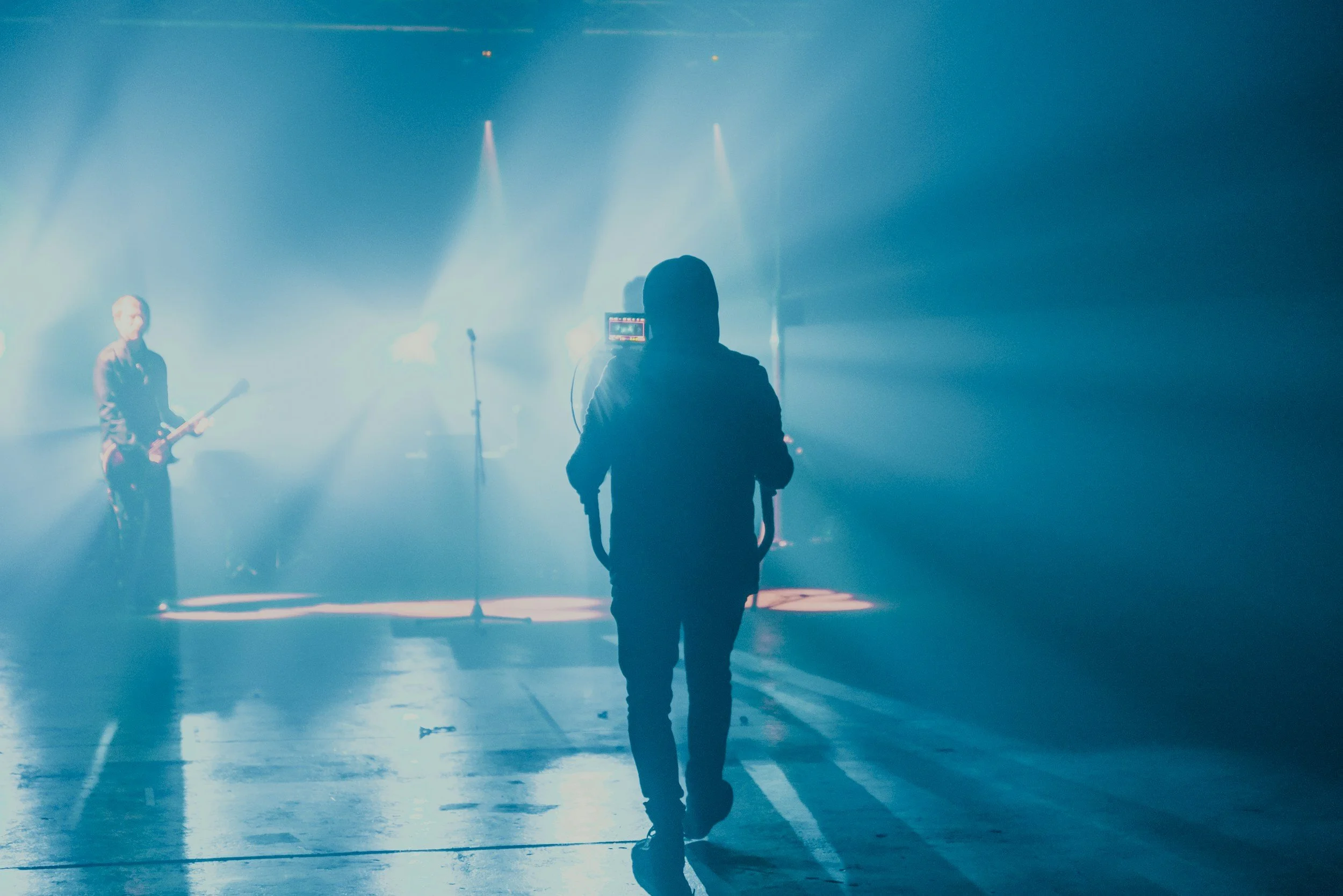 A person walking towards a stage where a band is performing, with blue stage lighting and a foggy atmosphere.