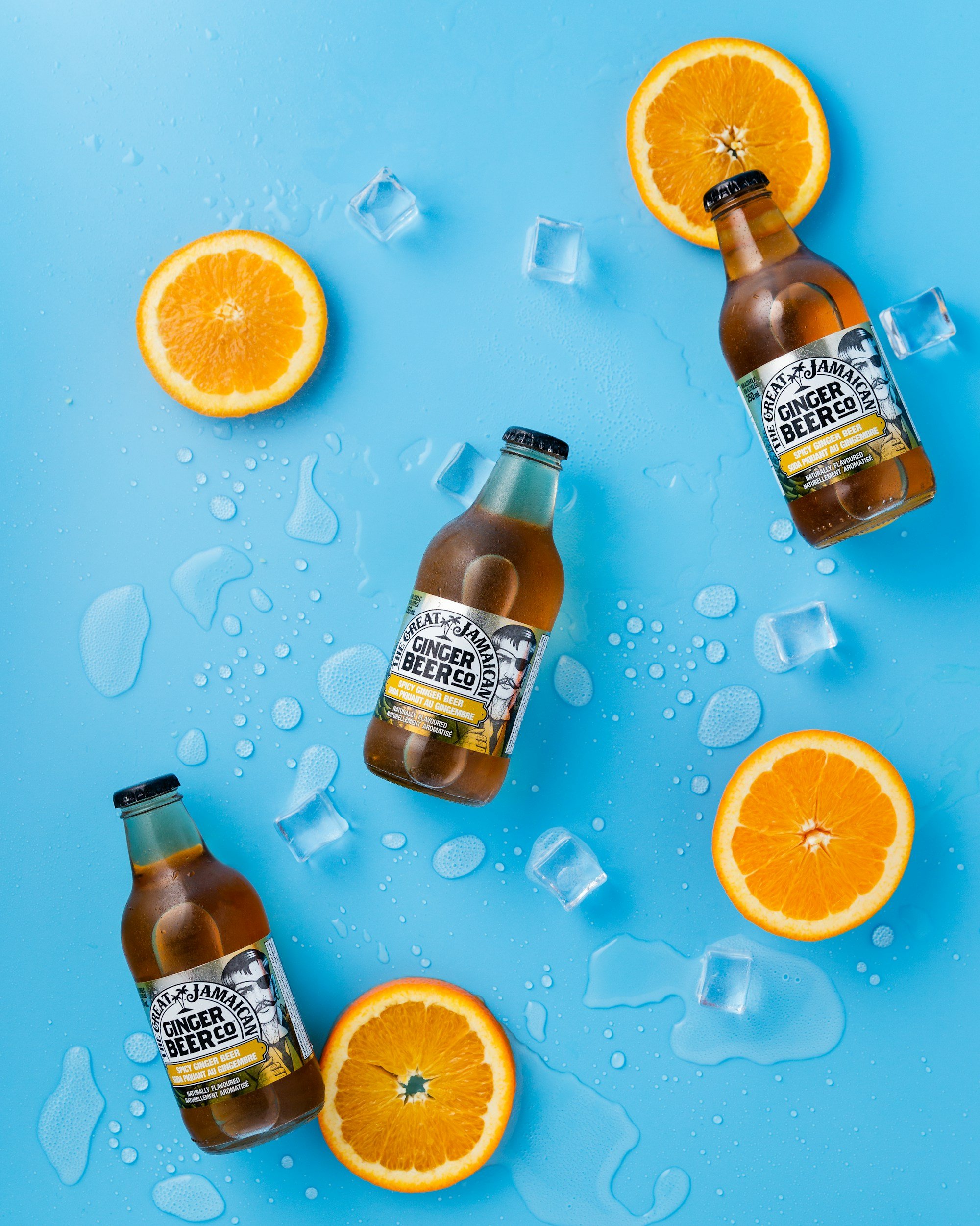 Three bottles of ginger beer with orange halves and ice cubes on a wet blue surface.