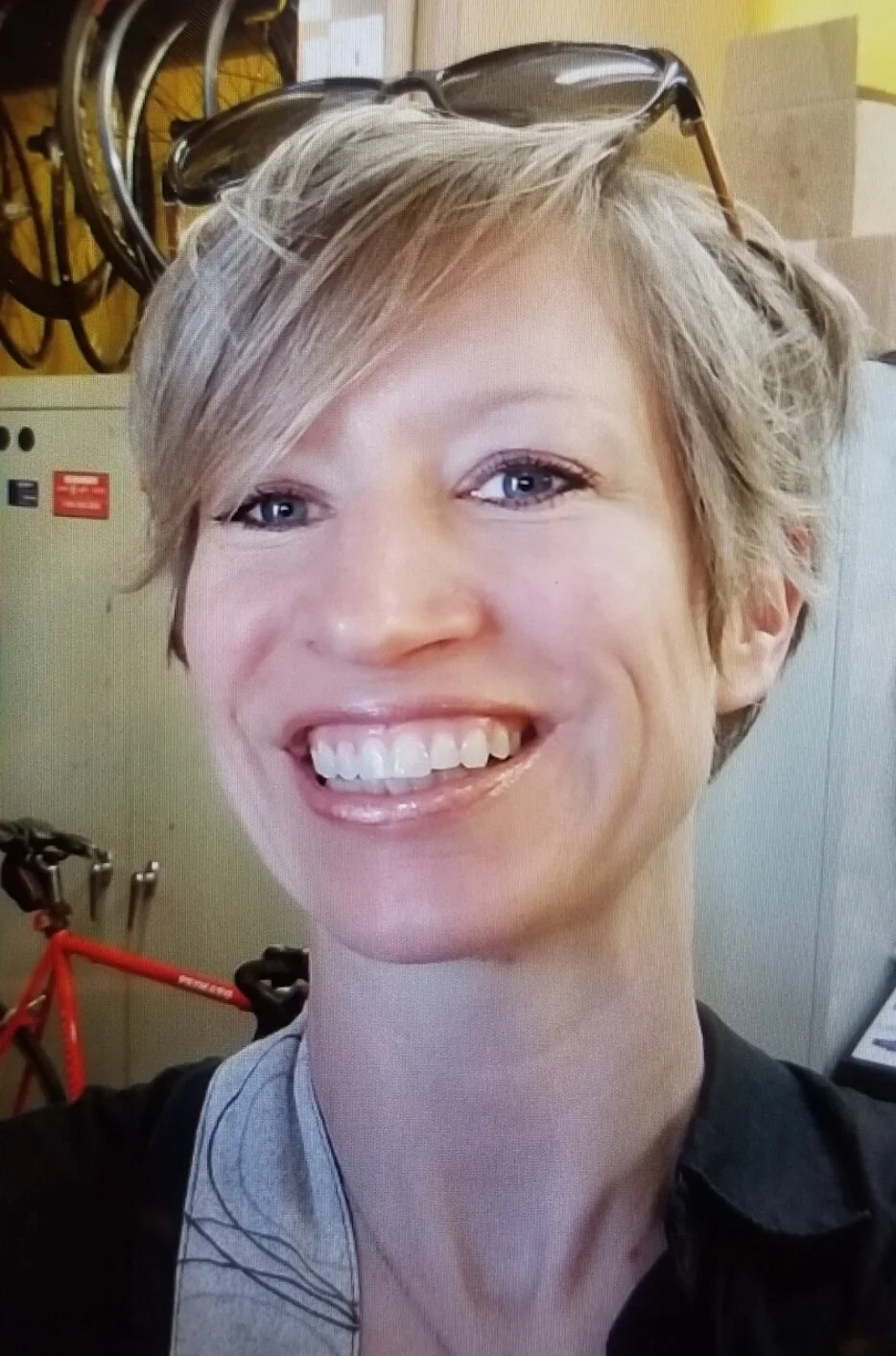 Close-up of smiling woman with short, blonde hair, glasses resting on her head, and a black shirt, with a red bicycle and bicycle helmets in the background.