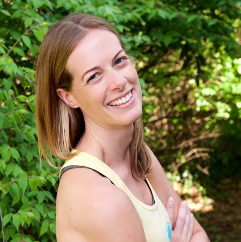 A smiling woman with shoulder-length light brown hair, wearing a yellow tank top, standing outdoors in front of green foliage.