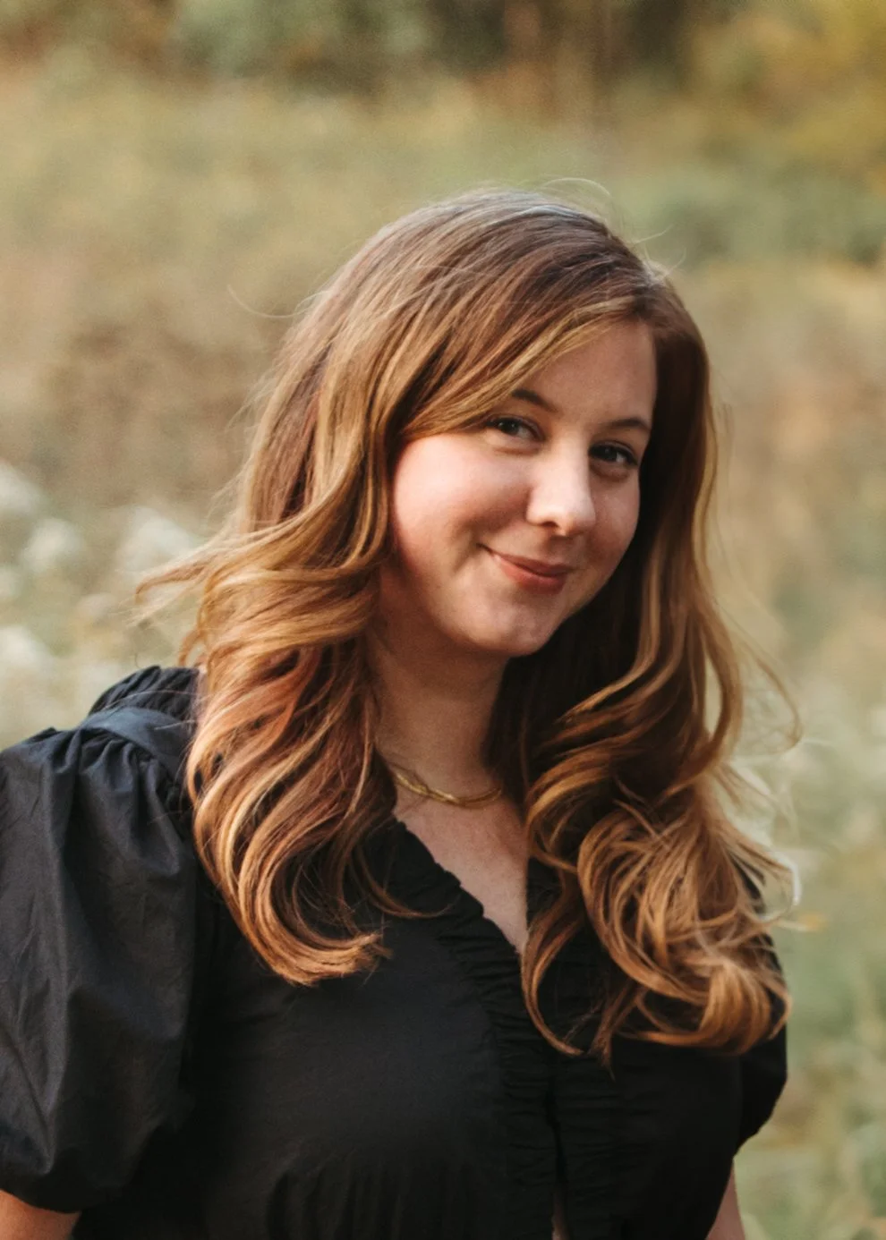 A young woman with long wavy reddish-brown hair smiling outdoors against a blurred natural background.