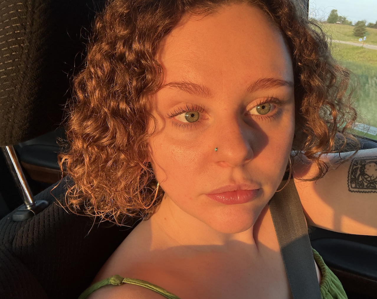 Close-up of a woman with curly hair, green eyes, and a nose piercing, sitting in a car during sunset with sunlight illuminating her face.