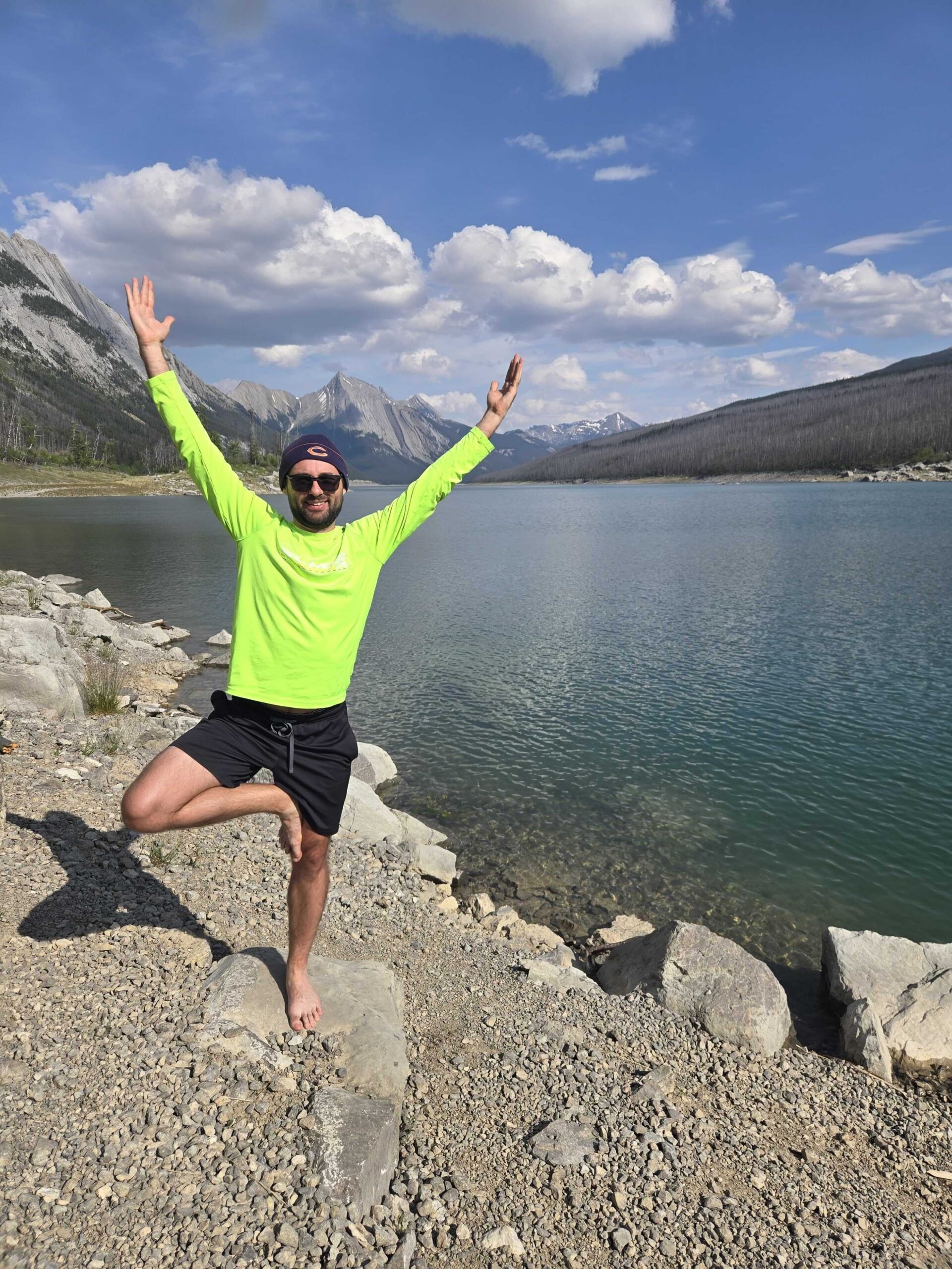 A man in a bright yellow shirt, black shorts, and sunglasses is standing on rocks by a lake, with mountains and a partly cloudy sky in the background, balancing on one foot with arms raised in a yoga pose.