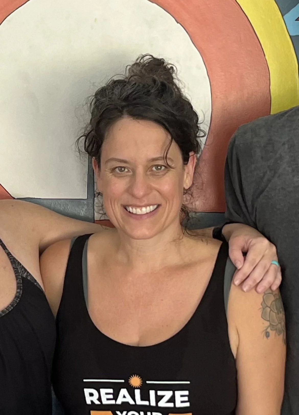 Smiling woman with short dark hair, wearing a black tank top that says "Realize Your Potential" in front of a colorful mural.