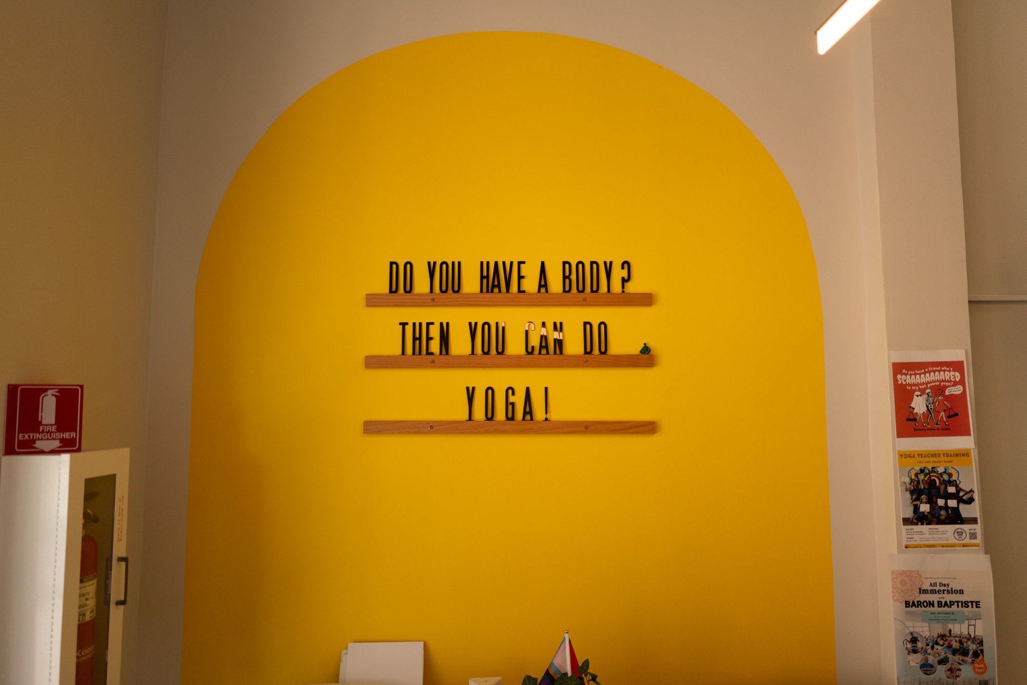 Yellow wall with black letters asking "Do you have a body? Then you can do yoga" on wooden shelves. Surrounding walls have various posters and a fire extinguisher.