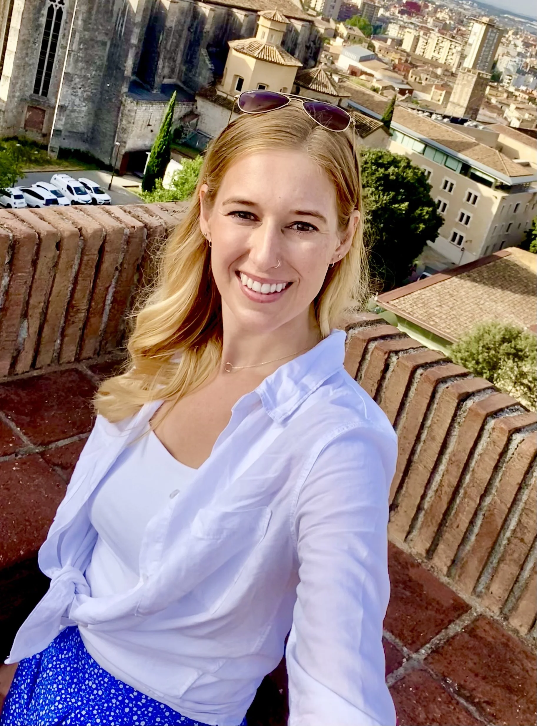 A woman with blonde hair and sunglasses on her head taking a selfie on a rooftop with a cityscape background.