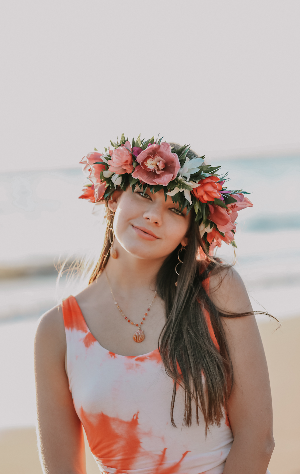 Young woman with long dark hair wearing a flower crown, a sleeveless white and orange tie-dye dress, and jewelry, standing on a beach during sunset.