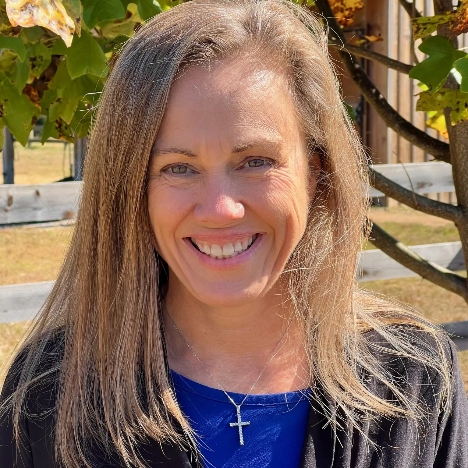 Smiling woman with long blonde hair and a cross necklace, outdoors with trees and a fence behind her.