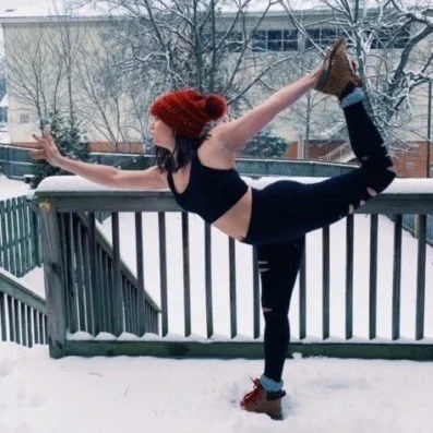 A woman practicing yoga outdoors in the snow, balancing on one leg while holding the other foot with her hand behind her back in a dancer pose, wearing a red knit hat, black sports bra, and black leggings.