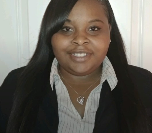 A woman with long black hair, wearing a black blazer over a striped dress shirt, smiling in front of a white background.