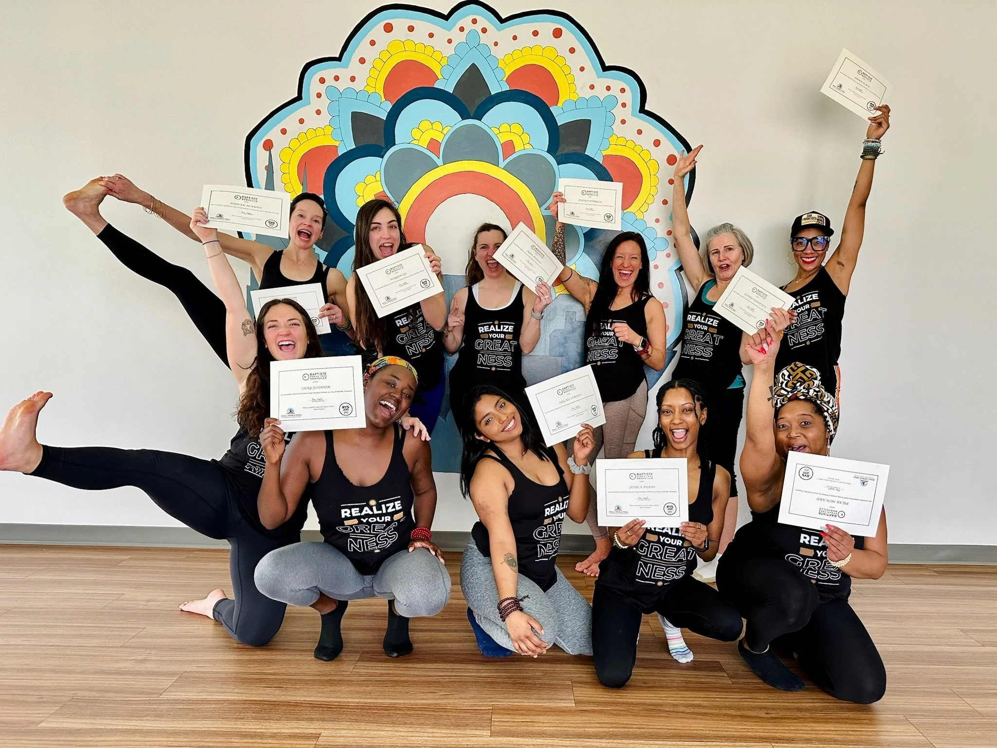 Group of women celebrating with certificates in front of a colorful mandala mural, some with arms raised and smiling.