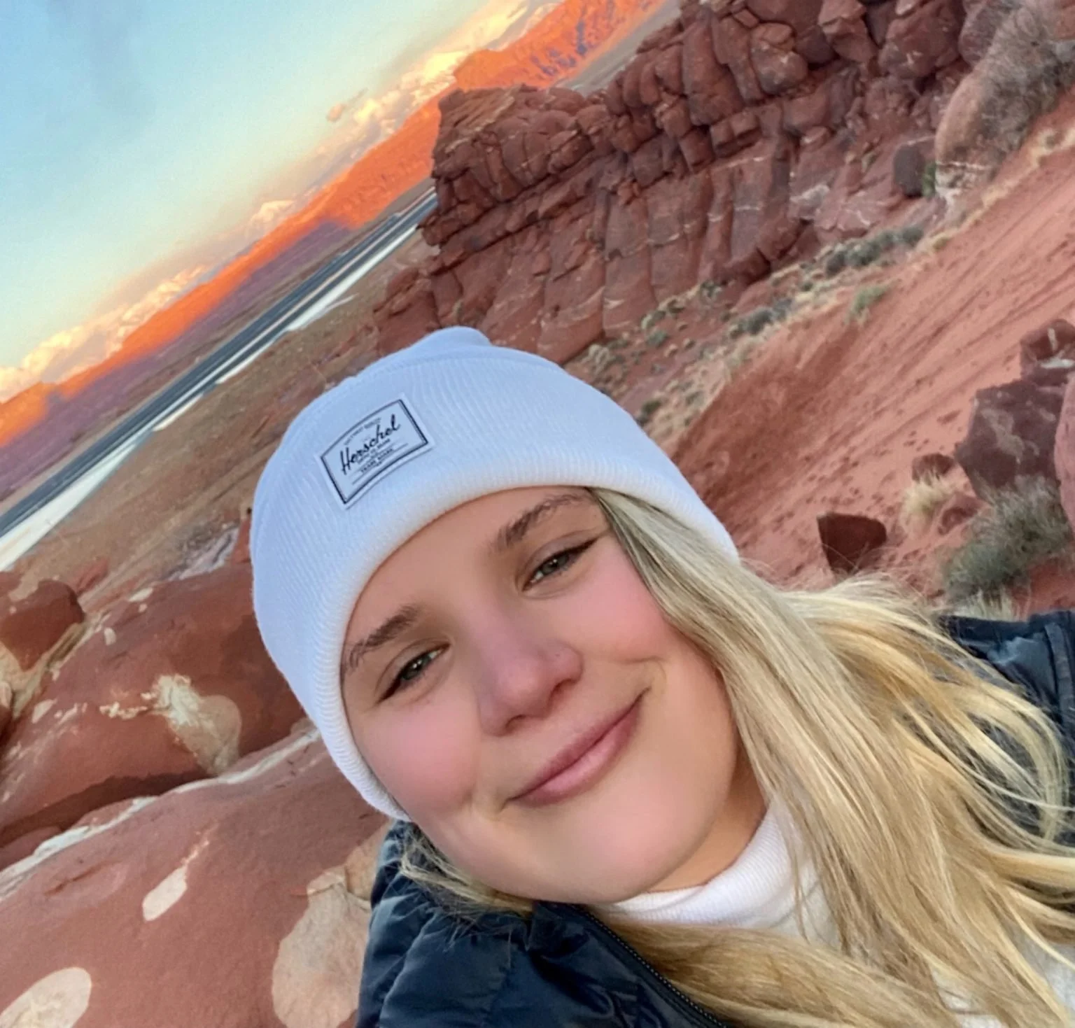 A smiling young woman with blonde hair wearing a white beanie hat and a black jacket, in a desert landscape with red rocks and distant mountains at sunset.
