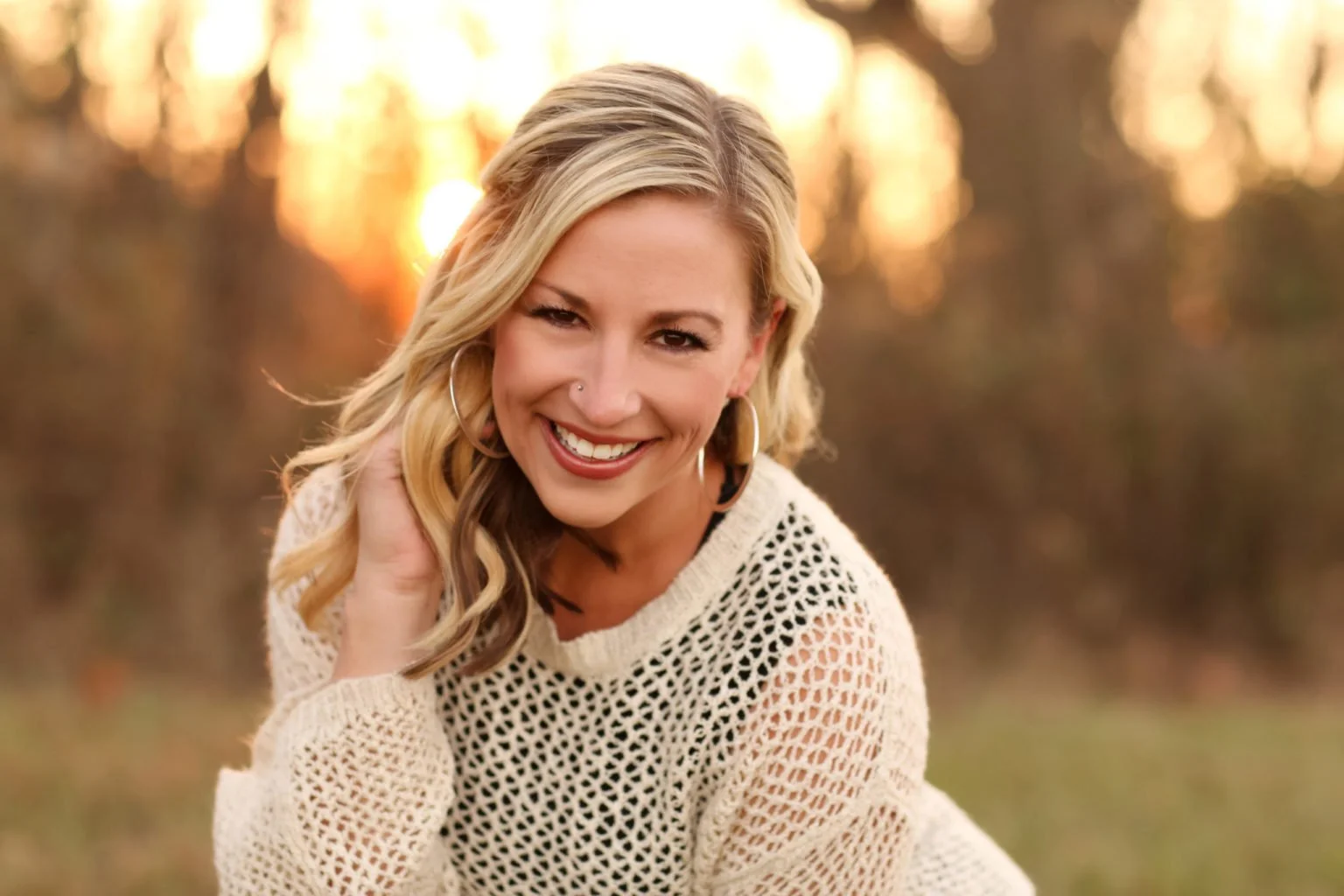 A smiling woman with blonde wavy hair, wearing hoop earrings and a cream-colored knitted sweater, sitting outdoors during sunset with a blurred background of trees and warm light
