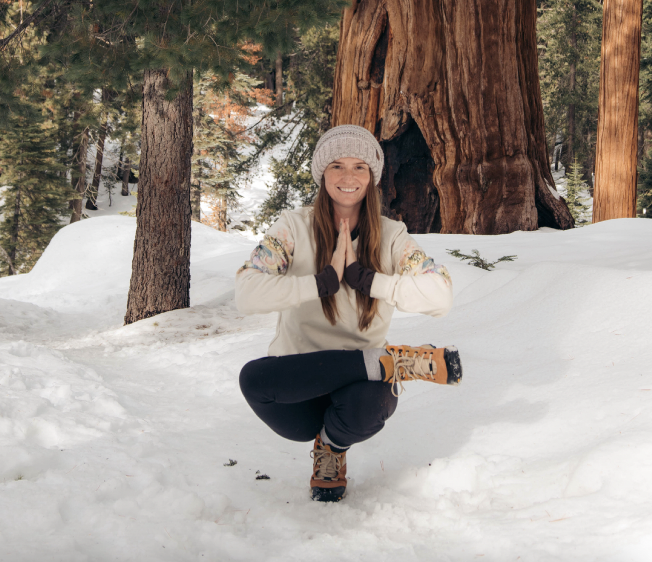 A woman in winter clothing poses with a yoga pose on the snow in a forest with tall trees and snow-covered ground.