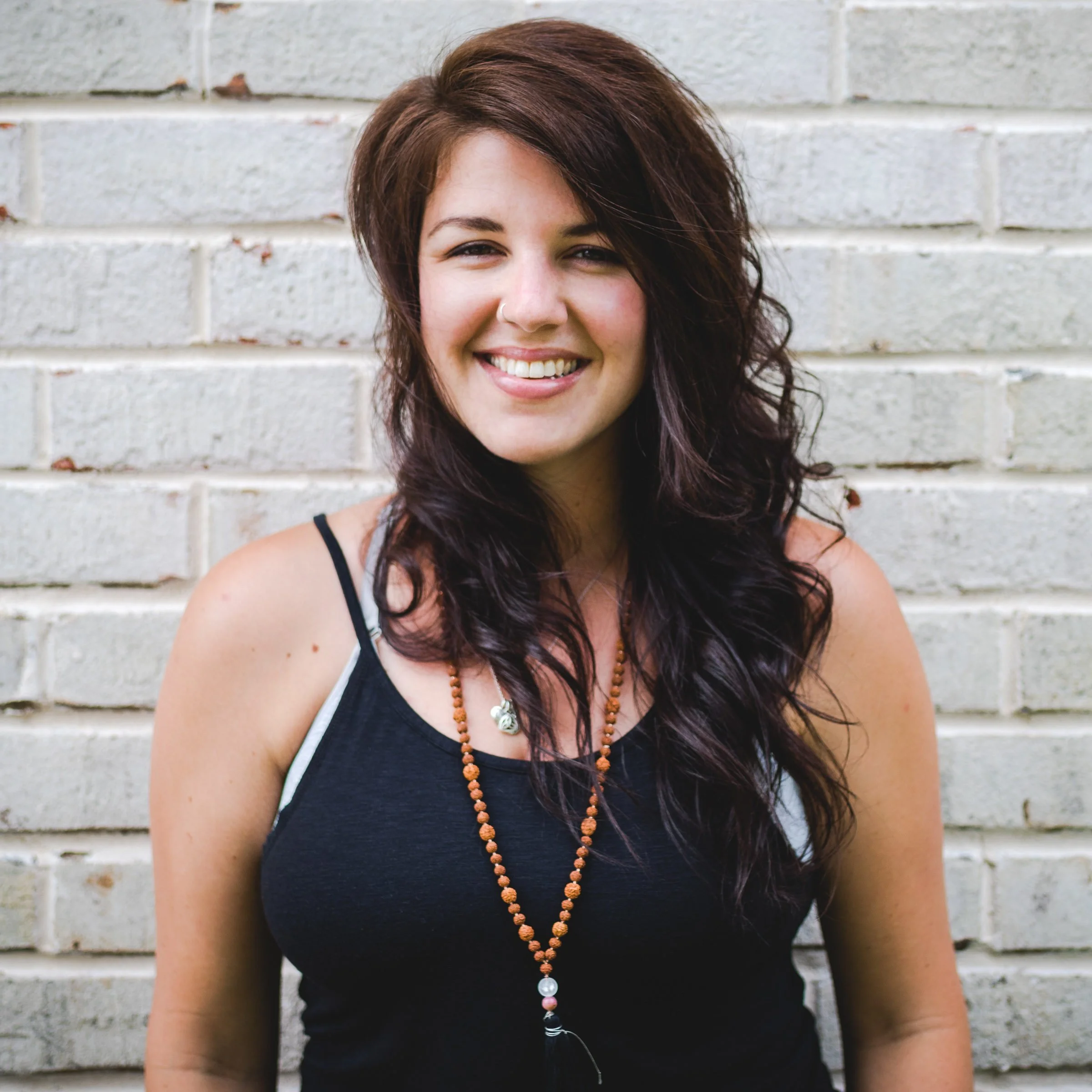 Smiling woman with long, wavy dark hair standing against a white brick wall.