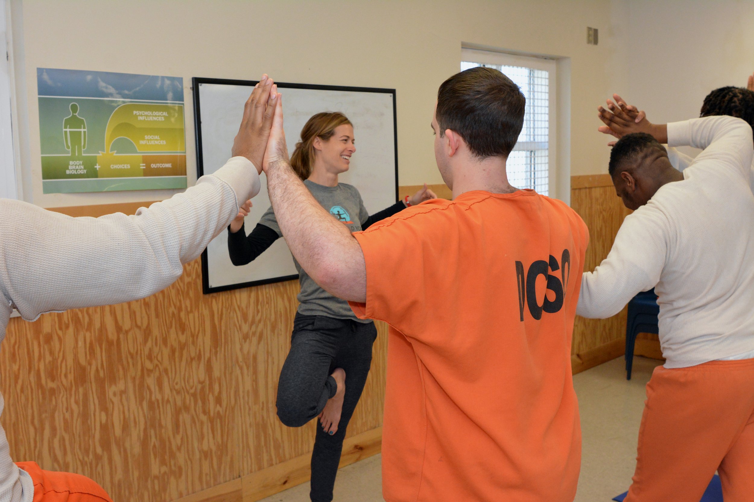 Group of people in a classroom doing a yoga pose with a woman instructor in front, who is smiling, holding a tree pose, and standing on one leg.
