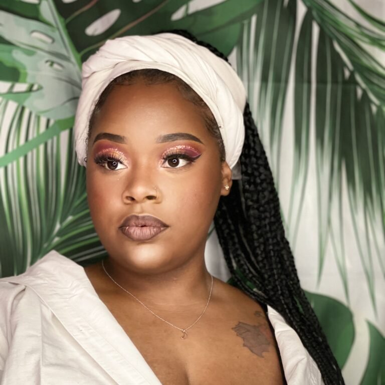 A young woman with dark skin and long braided hair, wearing a white headscarf and matching blouse, stands in front of a tropical leaf background with pink and gold eyeshadow, full lips, and a tattoo on her chest.