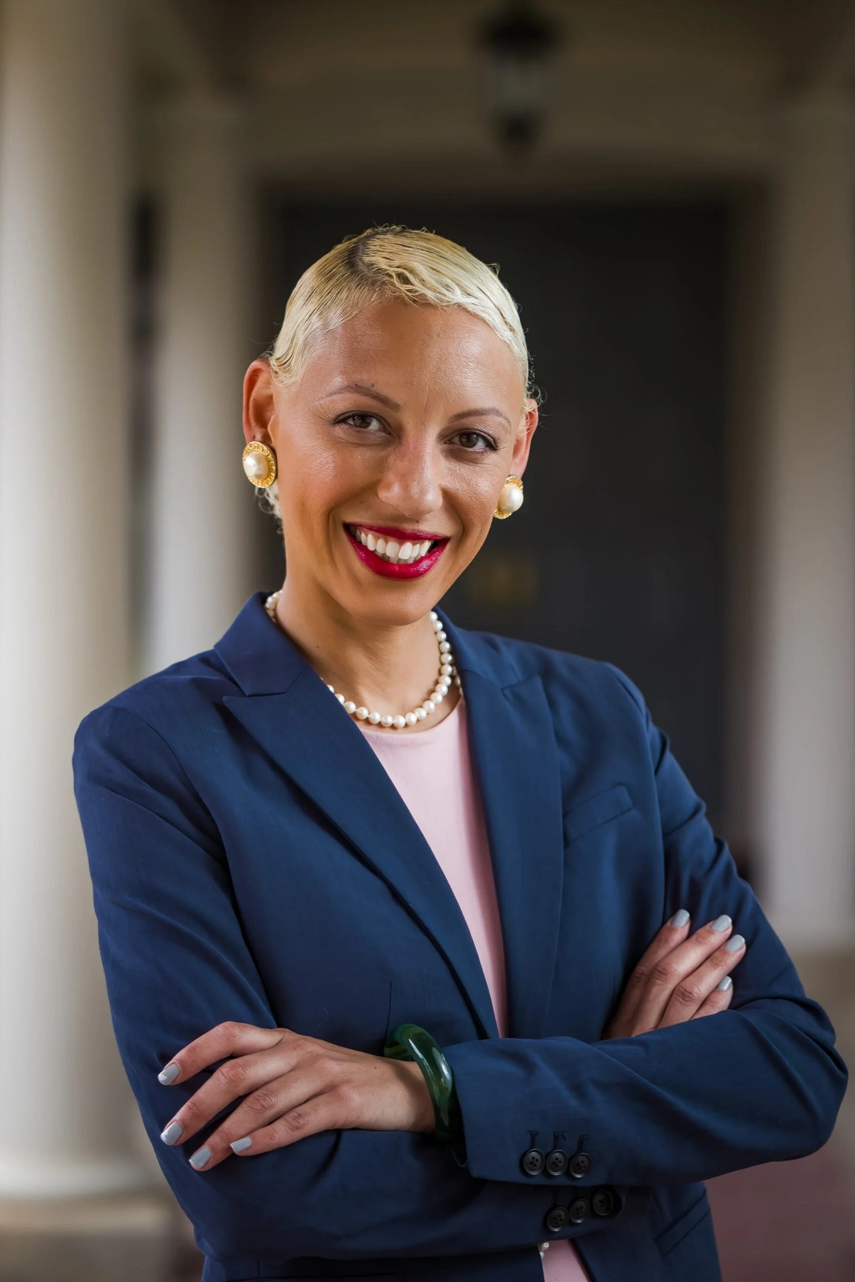 A confident woman with short platinum blonde hair, wearing a navy blazer, light pink blouse, pearl necklace, and pearl earrings, standing with arms crossed in a hallway.