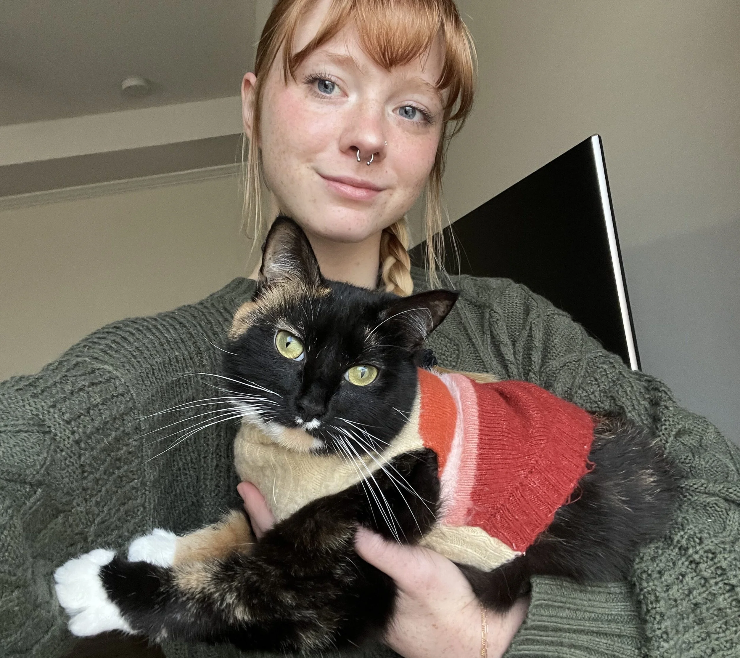 A young woman with red hair and freckles holding a black and tan cat wearing a sweater, indoors.