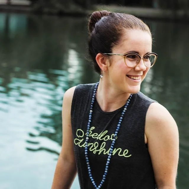 A smiling woman with glasses and a bun hairstyle, wearing a sleeveless black shirt with the words "Hello Sunshine," and a blue beaded necklace, standing outdoors near a body of water.
