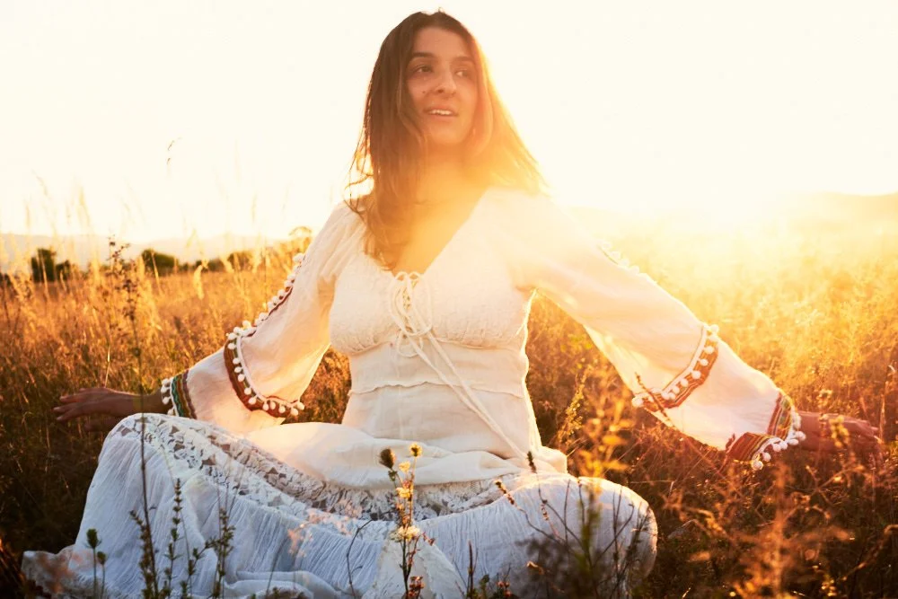 A woman sitting in a field of tall grass during sunset, wearing a white flowy dress with embroidered sleeves.