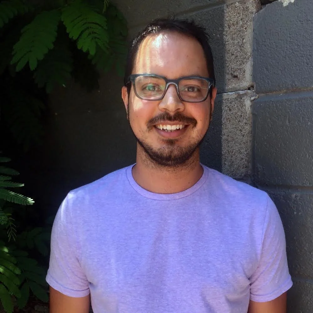 A smiling man with glasses and facial hair standing outdoors against a gray stone wall with green foliage in the background.