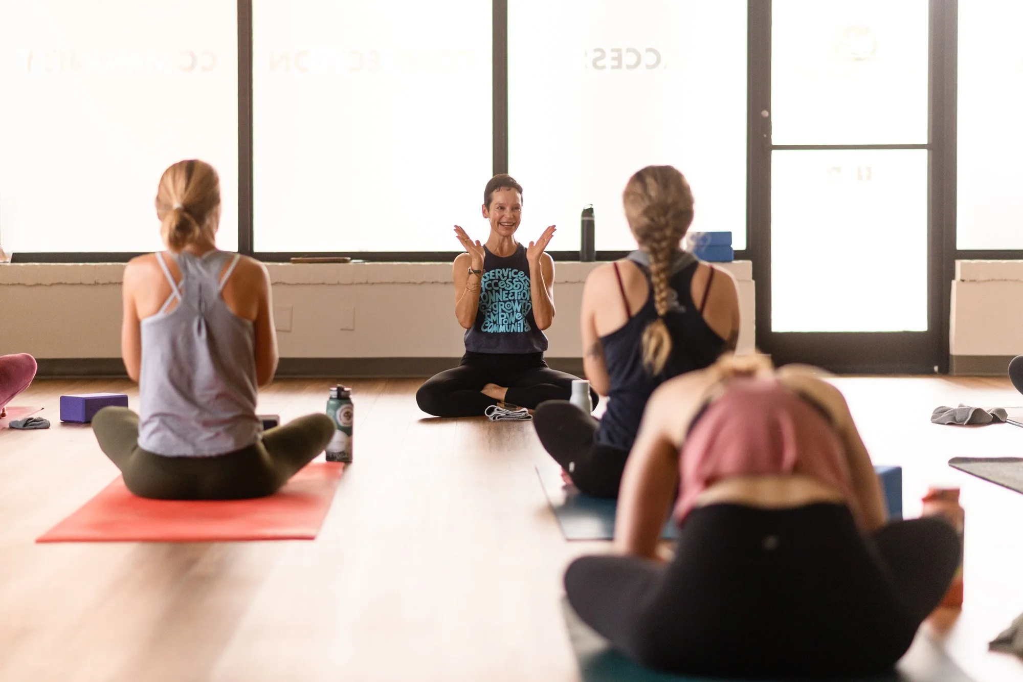 A woman instructor leading a yoga class with four women students sitting cross-legged on yoga mats, in a bright studio with large windows.
